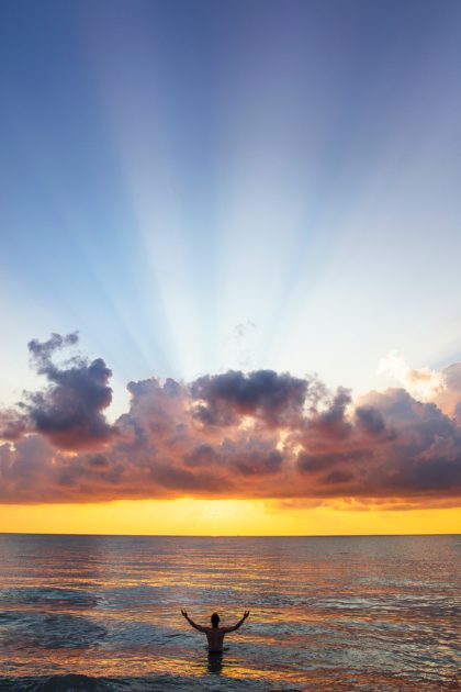 person, waist-deep in the ocean, arms outstretched, looking at dramatic sunrise with rays of light through the clouds