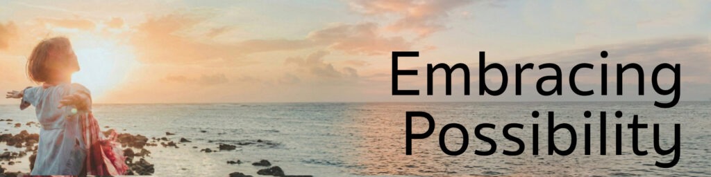 A white woman with short blonde hair wearing a denim jacket and red and white checked scarf is facing the ocean with the sun rising over the water. Her arms are spread wide. Text reads: Embracing Possibility